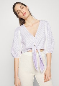 Blusa lilás cropped com decote em V e mangas bufantes, apresentando um padrão de diamantes, laço na frente e botões. Usada com calças texturizadas de cor clara.
