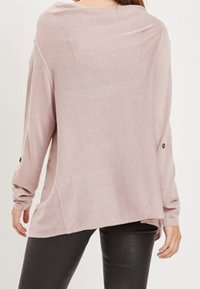 Femme portant un haut ample à manches longues rose clair avec des détails de boutons sur les manches et un pantalon noir ajusté, vue de dos.