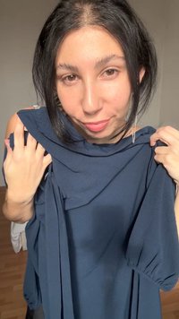 Tânără cu păr închis la culoare ține în mâini o vestimentație albastru închis, privind direct către camera cu un zâmbet subtil într-o cameră luminată difuz.