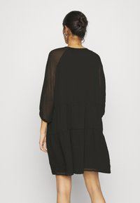 Vestido negro en capas, hecho de una tela liviana y texturizada, con mangas largas y transparentes y un escote redondo. La línea del dobladillo cae por encima de la rodilla.