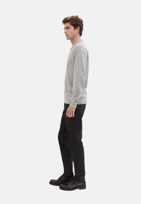 Grijze sweatshirt met lange mouwen, geribbelde manchetten en zoom, gecombineerd met zwarte broek en zwarte schoenen, zijaanzicht op een witte achtergrond.