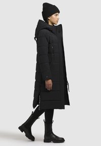 Cappotto lungo imbottito nero con cappuccio, tasche con zip e dettaglio con zip laterale. Indossato con un berretto lavorato a maglia nero e stivali alla caviglia.