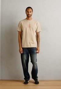 Beige pamut póló piros felirattal, sötétkék farmernadrággal párosítva. A modellen egy fából készült padlón áll, semleges fal előtt.