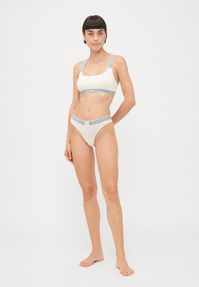 Beige bralette and matching thong featuring gray elastic bands with logo. Fabric is smooth; bralette has wide straps and a square neckline.