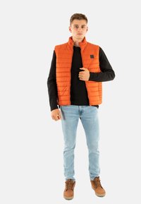 Oranje gewatteerde vest met een staande kraag, ritssluiting aan de voorkant en zwarte accenten, gecombineerd met een zwarte langemouwen shirt en lichtblauwe jeans.