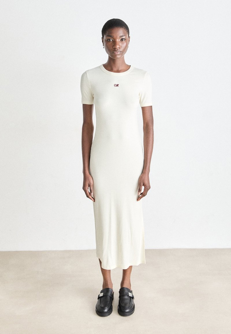 Knit midi dress in pale cream with short sleeves and a round neckline, featuring a small maroon logo. Worn with black slip-on shoes.