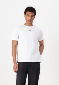 Hvid bomulds T-shirt med korte ærmer, der har et lille rundt logo på brystet. Pasformen er afslappet, og den er parret med mørke jeans.