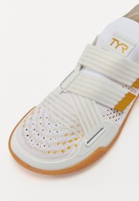 Λευκό και κίτρινο αθλητικό παπούτσι με αναπνεύσιμη πλεκτή επάνω επιφάνεια, δύο ευρείες ρυθμιζόμενες λωρίδες, καουτσούκ σόλα και λογότυπο TYR στη γλώσσα.