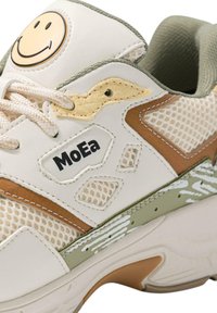 Hvite sneakers med nettingseksjoner, brune detaljer og en gul smilende ansiktlogo. Har teksturerte materialer og en gummisåle med grønne detaljer.