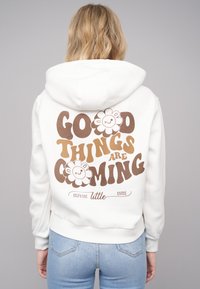 Hvit hettegenser med et stort brunt tekstdesign på ryggen som leser "GODE TING KOMMER" sammen med smilende blomsterillustrasjoner.