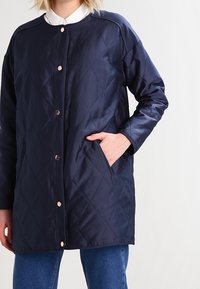 Osoba v prošívané námořnické modré bundě s patentkami, s jednou rukou v kapse, v kombinaci s modrými džíny a bílou košilí.