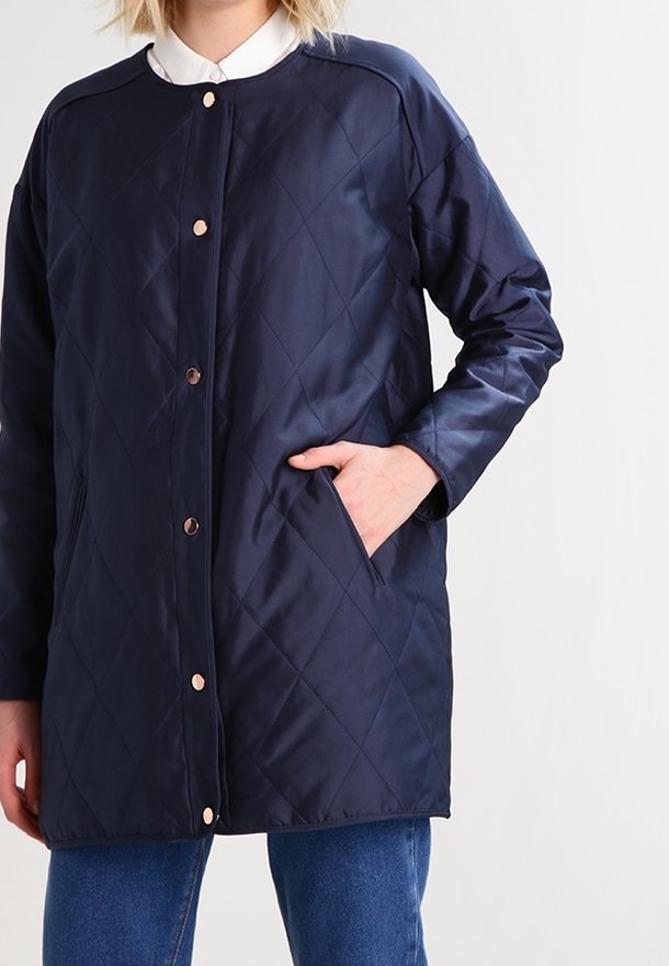 Osoba v prošívané námořnické modré bundě s patentkami, s jednou rukou v kapse, v kombinaci s modrými džíny a bílou košilí.