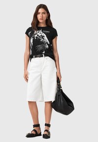 Camiseta negra de manga corta con gráfico de león, pantalones cortos blancos de talle alto, sandalias planas negras con tiras y un gran bolso negro.