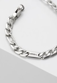 Pulsera de cadena de metal plateado con eslabones entrelazados, que presenta una textura suave y la inscripción "BREIL" en una placa pulida. Cierre de broche seguro.
