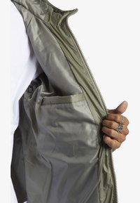 Olivgrön jacka med en glänsande, slät insida. Har en sidoficka, silverdragkedja och detaljerad krage med åtdragning.