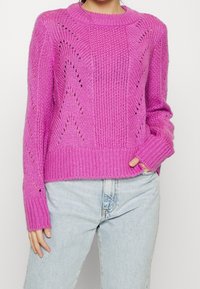 Kvinna som bär en ljusrosa stickad tröja med texturmönster och ljusblå högmidjade jeans, med handen i fickan, mot en enkel bakgrund.