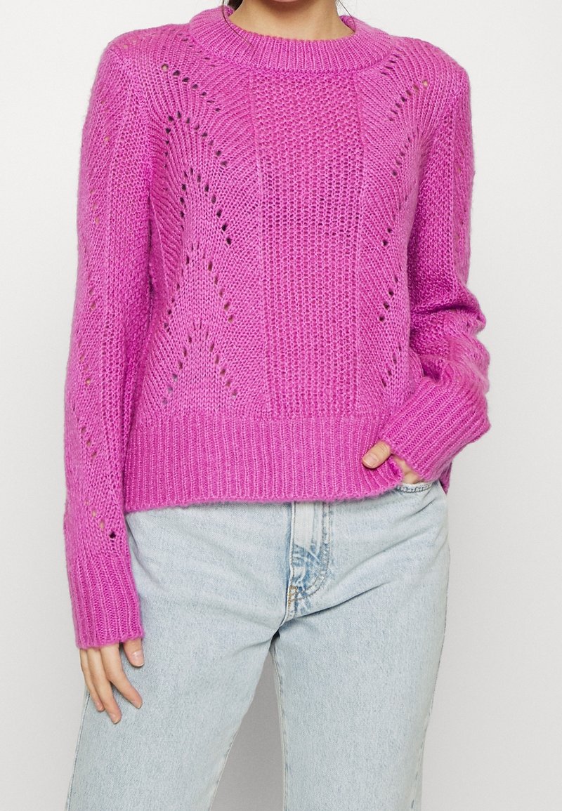 Kvinna som bär en ljusrosa stickad tröja med texturmönster och ljusblå högmidjade jeans, med handen i fickan, mot en enkel bakgrund.