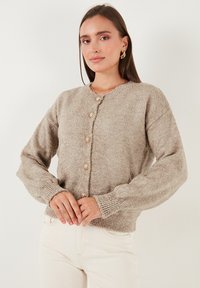 Bežinė megzta kardiganas su tekstūruotu audiniu, apvalia iškirpte, mygtukų uždarymu ir pundomais rankogaliais. Yra dideli dekoratyviniai mygtukai.