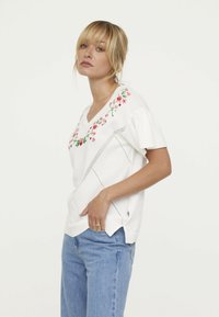 Top blanc à manches courtes avec broderies florales en rose et vert, avec un décolleté en V et un design texturé. Détails découpés le long des coutures.