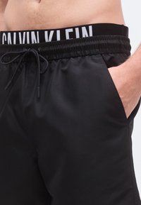 Μαύρο μαγιό σορτς με ελαστική μέση που φέρει το λογότυπο "CALVIN KLEIN" σε λευκό, ρυθμιζόμενο κορδόνι και πλαϊνές  τσέπες. Λείος υφής.