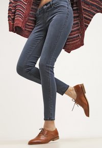 Jeans ajustados de color azul oscuro combinados con zapatos de cuero marrón con cordones. El suéter es rojo con rayas horizontales multicolores.
