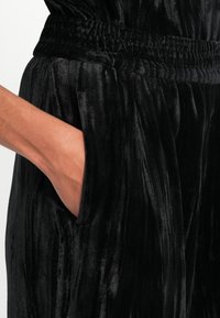 Main reposant dans la poche d'un pantalon en velours noir avec une taille élastique froncée et un tissu à texture douce.