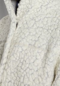 Chaqueta de forro polar blanca con una superficie texturizada y esponjosa, que presenta un cierre de cremallera y un detalle de costura visible a lo largo de la costura.