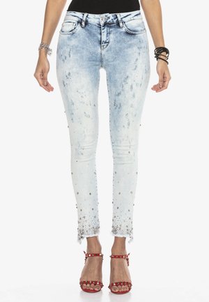 Kobieta ubrana w jasnoniebieskie obcisłe dżinsy z cyrkoniami i postrzępionymi brzegami, w połączeniu z czerwonymi sandałami na wysokim obcasie z ćwiekami oraz bransoletkami.