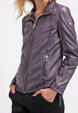 Nainen pukeutuneena istuvaan tummanruskeaan nahkatakkiin, jossa on vetoketju edessä ja taskut, kädet mustien housujen taskuissa, seisoo valkoisen taustan edessä.