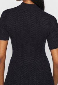 Abito maglione in maglia blu navy con un motivo a spina di pesce testurizzato, maniche corte e colletto alto. Il materiale sembra morbido e aderente.