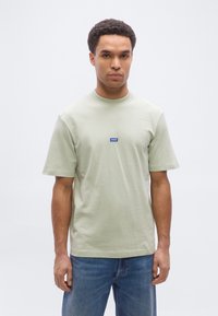 Άνδρας που φοράει ανοιχτοπράσινο κοντομάνικο μπλουζάκι με μικρό μπλε λογότυπο «HUGO» στο στήθος και μπλε τζιν, στέκεται μπροστά σε απλό λευκό φόντο.