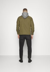 Hombre de pie frente a una pared, vistiendo una chaqueta verde oliva sobre una sudadera gris, jeans negros y zapatos negros, sobre un fondo blanco liso.