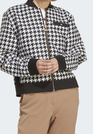 Personne portant une veste zippée à motif pied-de-poule noir et blanc avec des poignets noirs et un pantalon beige, les mains croisées devant.