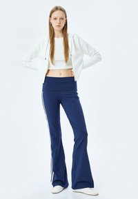 Witte cropped hoodie en marineblauwe flared broek met witte zijkanten, gemaakt van een gladde stof; model poseert tegen een effen achtergrond.