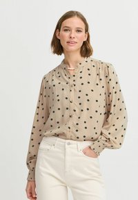 Beige halenka s černými puntíky, zapínání na knoflíky vpředu, vysoký límec a nařasené rukávy. Má pružné manžety a je vyrobena z měkké, lehké látky.