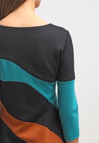 Haut noir à manches trois-quarts avec des blocs de couleur ondulés en bleu sarcelle et en orange brûlé, mettant en avant des textures contrastées et un tissu lisse.