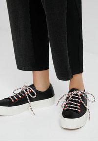 Baskets en cuir noir avec semelle plateforme blanche, lacets à motifs colorés et œillets métalliques. Portées avec un pantalon noir court.