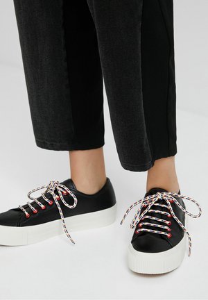 Baskets en cuir noir avec semelle plateforme blanche, lacets à motifs colorés et œillets métalliques. Portées avec un pantalon noir court.