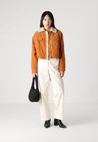 Veste en velours côtelé orange raccourcie avec un col en fausse fourrure beige, assortie à un pantalon blanc à jambes larges et un sac en cuir tressé noir.