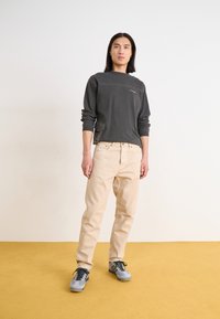 Maglia grigia a maniche lunghe con tasca, pantaloni beige e sneaker grigi. Tessuto morbido e vestibilità rilassata, con dettagli di cucitura visibili.