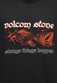 Μαύρο ύφασμα με πορτοκαλί αφηρημένο σχέδιο από κλήμα και φύλλα, λευκό κείμενο «volcom stone» πάνω και «strange things happen» κάτω από το σχέδιο.
