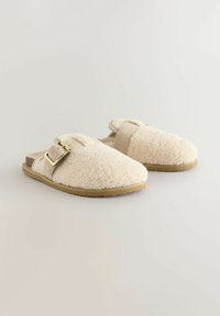 Chaussons en fourrure de couleur crème avec une surface texturée, strap réglable et boucle métallique sur une semelle en caoutchouc beige et flexible.