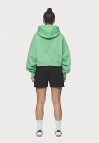 Sudadera verde recortada con capucha, mangas anchas, usada con pantalones cortos negros y calcetines blancos con acentos verdes, vista desde la parte de atrás.