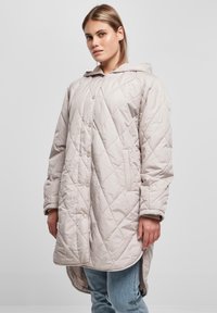 Quilted, lang, lys beige frakk med hette, med trykknapper og sidelommer, laget av glatt, myk tekstil.
