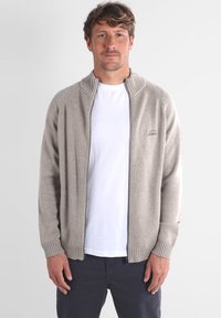 Cardigan zippé gris clair en tissu tricoté, avec un col haut et des poignets côtelés, porté par-dessus un t-shirt blanc.