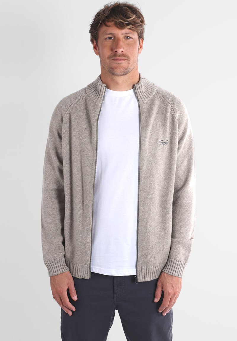 Cardigan zippé gris clair en tissu tricoté, avec un col haut et des poignets côtelés, porté par-dessus un t-shirt blanc.