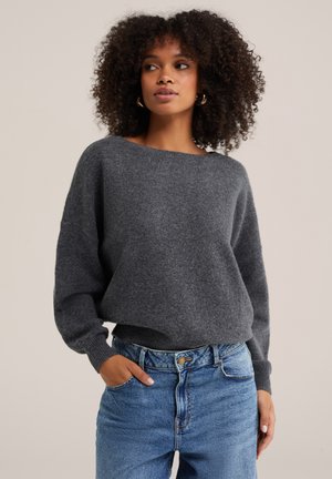 Kvinde med krøllet hår iført guldringeøreringe, en mørkegrå sweater og blå cowboybukser med høj talje, med den ene hånd i lommen.