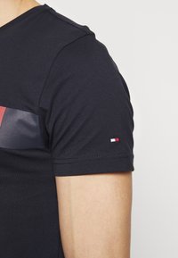 Close-up of a person’s arm wearing a black short-sleeve shirt with a small red, white, and blue logo on the sleeve.