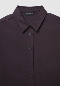 Camicia button-up di un viola scuro con colletto classico, cinque bottoni neri e una superficie liscia e tessuta. Etichetta del marchio "CALLIOPE" all'interno del colletto.