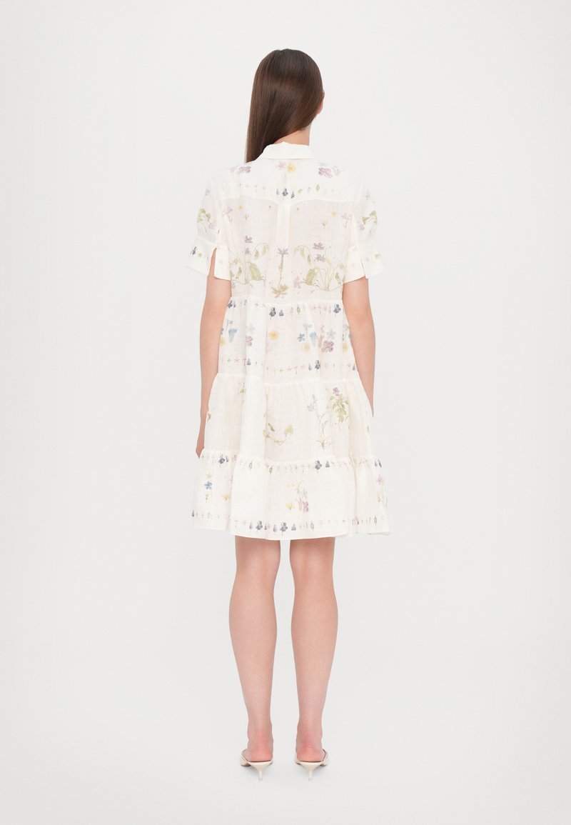 Robe en lin blanc avec des manches courtes, un design à volants et des broderies florales dans des couleurs pastel. Présente un col et une fermeture à boutons au dos.